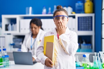 Mother and young daughter working at scientist laboratory covering mouth with hand, shocked and...