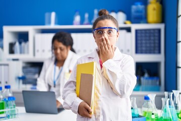 Mother and young daughter working at scientist laboratory covering mouth with hand, shocked and...