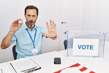 Middle age man with beard sitting by ballot holding i vote badge doing stop gesture with hands palms, angry and frustration expression