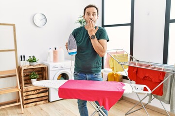 Middle age man with beard ironing clothes at home covering mouth with hand, shocked and afraid for mistake. surprised expression