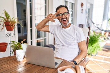 Middle age man using computer laptop at home doing peace symbol with fingers over face, smiling...