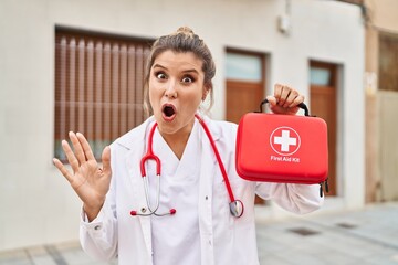 Young doctor woman holding first aid kit scared and amazed with open mouth for surprise, disbelief...