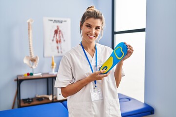 Young woman holding shoe insole at physiotherapy clinic smiling with a happy and cool smile on face. showing teeth. - obrazy, fototapety, plakaty
