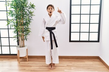 Young hispanic girl wearing karate kimono and black belt pointing finger up with successful idea. exited and happy. number one.