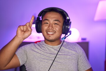 Chinese young man playing video games wearing headphones smiling with an idea or question pointing...