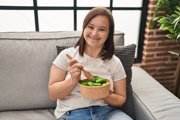 Down syndrome woman eating salad sitting on sofa at home