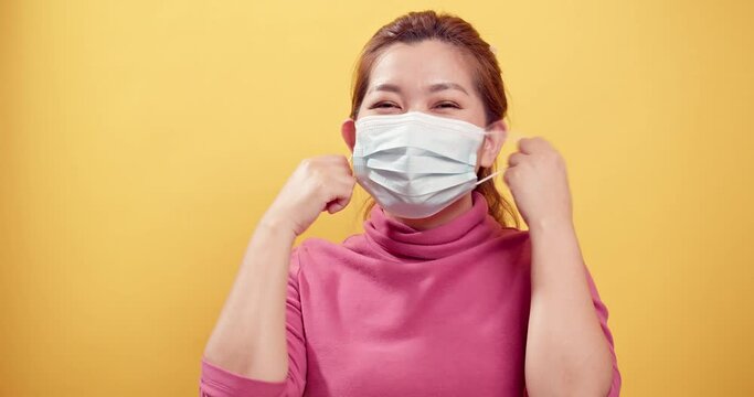 Asian gorgeous young woman smiling and taking off the protective mask for fresh air after the CIVID-19 situation unravels with the yellow background.