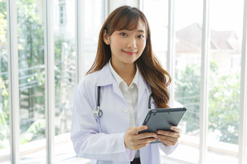 Beautiful and cheerful good mood young lady asian doctor dressed uniform smiling arm stethoscope while using tablet on lobby hospital background...