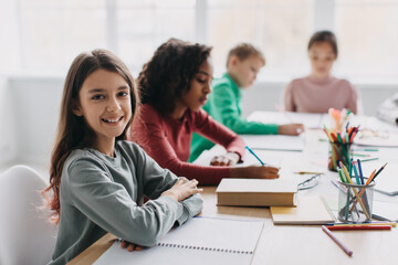 Happy Schoolgirl Sitting At Desk Smiling To Camera In Classroom