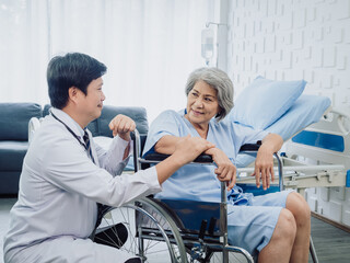 Obraz premium Asian senior woman patient dressed in blue, smiling happily sits in wheelchair with kindly man doctor in white suit holding her hand beside in recovery room in hospital. Healthcare support concept.