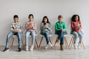 Group Of Amazed Diverse School Kids Using Smartphones Sitting Indoors