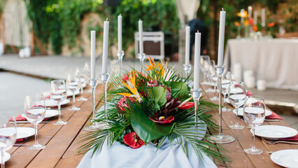 Wedding banquet decor in tropical style. In the center of the table is a white tablecloth, a bouquet of strelitzia, red anthurium, and tropical leaves. On the sides are candles, wine glasses, plates.