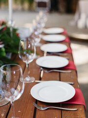 Decor and serving of a wedding banquet on a wooden table. White plates on a red napkin, cutlery, and wine glasses. Tropical flowers in the background.