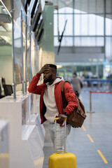 Upset depressed African American man with luggage standing at check-in counter in airport, being...