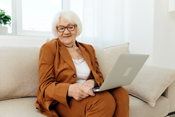 a joyful elderly woman is mastering new technologies using the Internet on a laptop while sitting on the couch at home