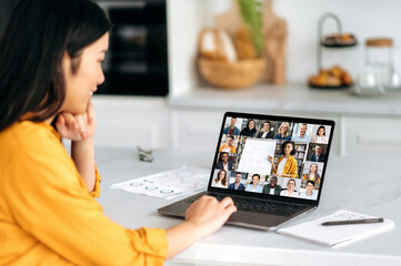 Obraz na płótnie Canvas Remote lesson. Positive smart Asian female student, sitting at the table, listening to an online lecture, on the screen of a laptop, a teacher telling information, and a group of multiracial people