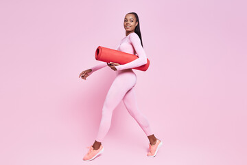 Beautiful African woman carrying exercise mat while walking against pink background