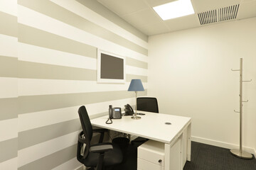 Office cubicle with small white double table with telephones, black swivel chairs, metal coat rack, scratched wall and dark gray carpet floor