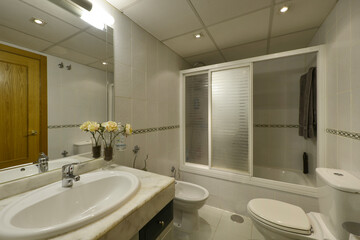 Bathroom with white porcelain sink with marble top on the chest of drawers, frameless mirror on the wall and shower cabin with sliding screen with white aluminum edges
