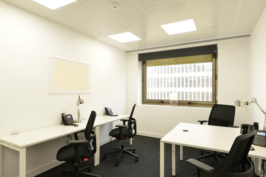 Office cubicle with a small white double table with telephones, black swivel chairs, a window with a view and a dark gray carpet floor