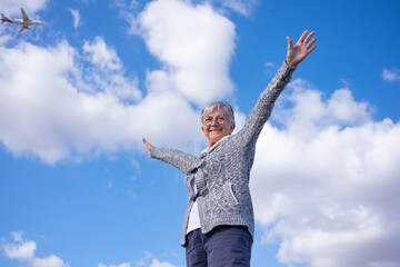 Smiling senior woman outdoors with outstretched arms in the blue sky while a plane passes in the sky.