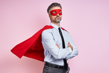 Confident man in shirt and tie wearing superhero cape and keeping arms crossed against pink...
