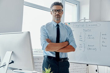 Confident English teacher looking at camera while standing near the whiteboard at classroom