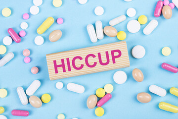 On a blue background, multi-colored pills and a wooden block with the text HICCUP. View from above....