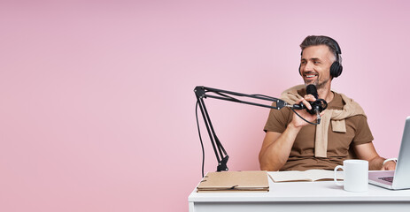 Cheerful man in headphones using microphone while recording podcast against pink background