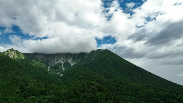Flying over green beech forest on slopes leading to clouded mountain peak