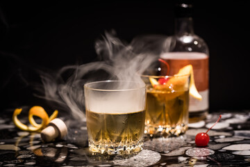Smoke billowing out of an Old Fashioned cocktail, against a black background.