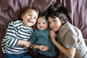 Portrait of two brothers lying on bed with their baby girl sibling, positive children of different...