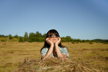 Naklejka na ściany i meble A beautiful girl in a summer dress and sunglasses near a haystack enjoys her vacation.