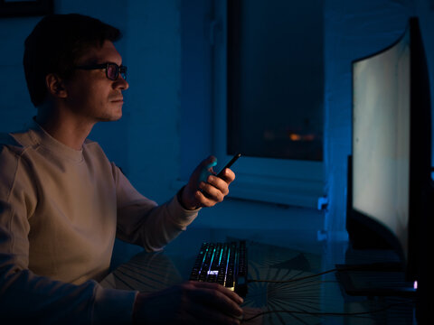 Programming and development of cross-platform applications, a young man with glasses is sitting at a computer and working on an online project. Dark room, shallow depth of field