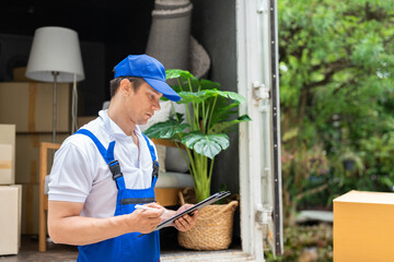 Man mover worker in blue uniform checking lists on clipboard while unloading cardboard boxes from truck.Professional delivery and moving service.