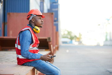 African factory worker or engineer holding smartphone and looking to something in containers...