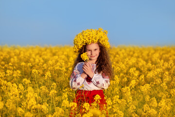 Naklejka na ściany i meble Ukrainian child girl in traditional embroidery and yellow wreath in field of yellow flowers against blue sky. Pray for Ukraine. Ukraine's Independence Flag Day. Constitution day. Symbols of Ukraine.