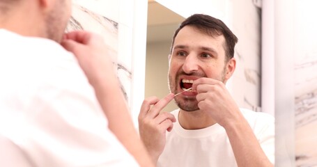 Health care, dental hygiene, people and beauty concept - smiling young man with floss cleaning teeth and looking to mirror at home bathroom.