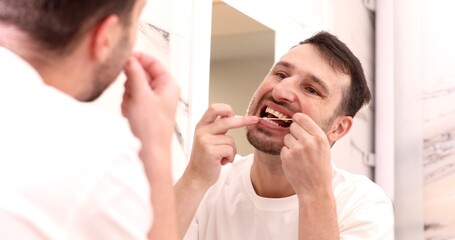 Health care, dental hygiene, people and beauty concept - smiling young man with floss cleaning teeth and looking to mirror at home bathroom.