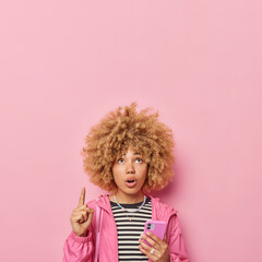 Shocked embarrassed young woman with curly hair points index finger above cannot believe in amazing news holds mobile phone scrolls social networks dressed in striped jumper and windbreaker.