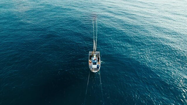 White small fisherman boat with people on board waving in ocean. People work together to pull out trawl with caught fish from deep blue waters. Aerial frontal shot with object in center
