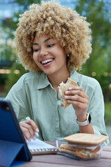 Happy curly haired young woman uses tablet and takes notes in notebook creats article laughs gladfully as watches funny video wears shirt smartwatch poses at wooden table on blurred background