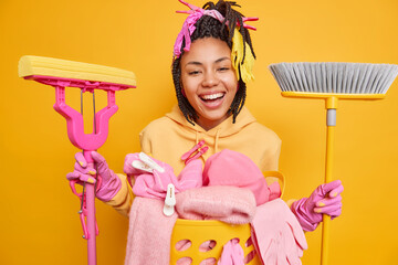 Cheerful young woman with braided hair holds mop and broom smiles happily wears sweatshirt and...