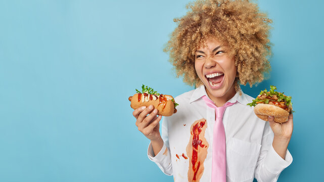 Horizontal shot of emotional curly haired woman eats fast food has unhealthy nutrition habits exclaims loudly dressed in formal outfit poses against blue background blank space on left for promotion