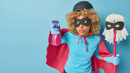 Horizontal shot of serious curly haired woman pretends being superhero holds spray detergent and mop ready for cleaning house isolated over blue background blank space for your advertisement