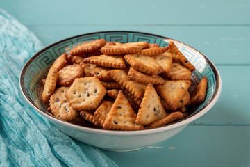 Teal bowl full of saltine crackers over turquoise wood background. Salty crackers with black and white sesame seeds in a blue deep plate. Ready to eat baked crunchy snack. Saltines concept.