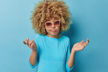Concerned hesitant woman with blonde curly hair spreads palms feels unaware and hesitant wears...