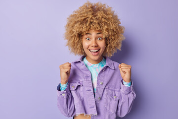 Indoor shot of emotional curly haired woman clenches fists feels excited shouts for favorite team while watching football match wears stylish jacket poses over purple background celebrates something