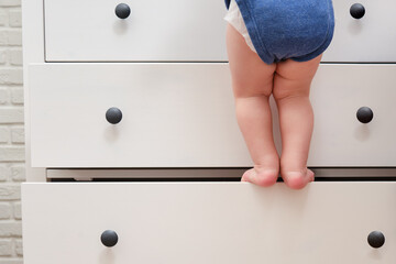 Toddler baby climbed up on the open chest of drawers. Child boy stood on a tall drawer of a white...