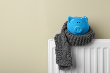 Piggy bank wrapped in scarf on heating radiator against beige background, space for text